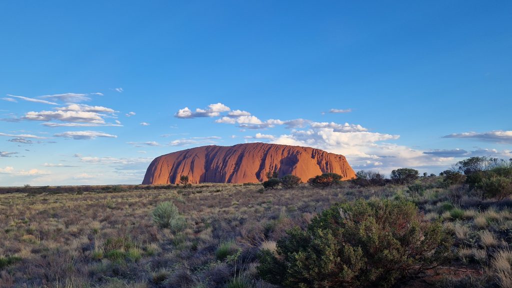 Der riesige rote Berg Uluru leuchtet in den letzten Sonnenstrahlen umgeben von der kargen Landschaft des Outbacks in Australien