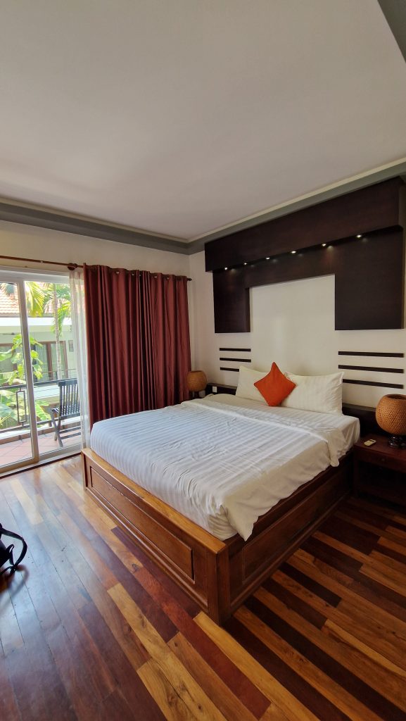 Ein Zimmer im Hotel Sakanban Suite in Siam Reap. Licht fällt vom Balkon in den hellen raum mit Holzpaket. Das Bett steht in der Mitte des Raums. Das Zimmer ist geräumig.