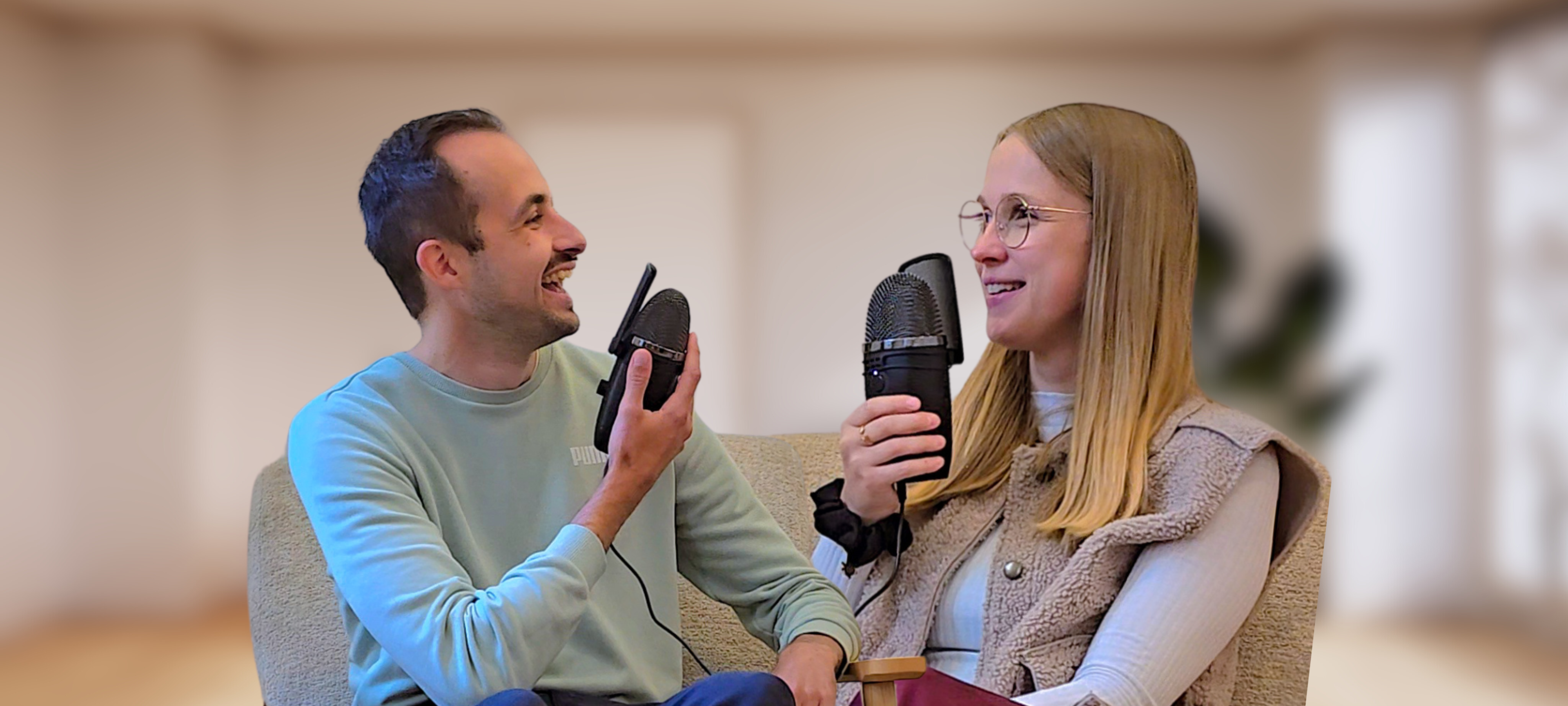 Lena und Philippe sitzen sich mit Mikrophonen in den Händen auf Sesseln gegenüber und lachen sich an.