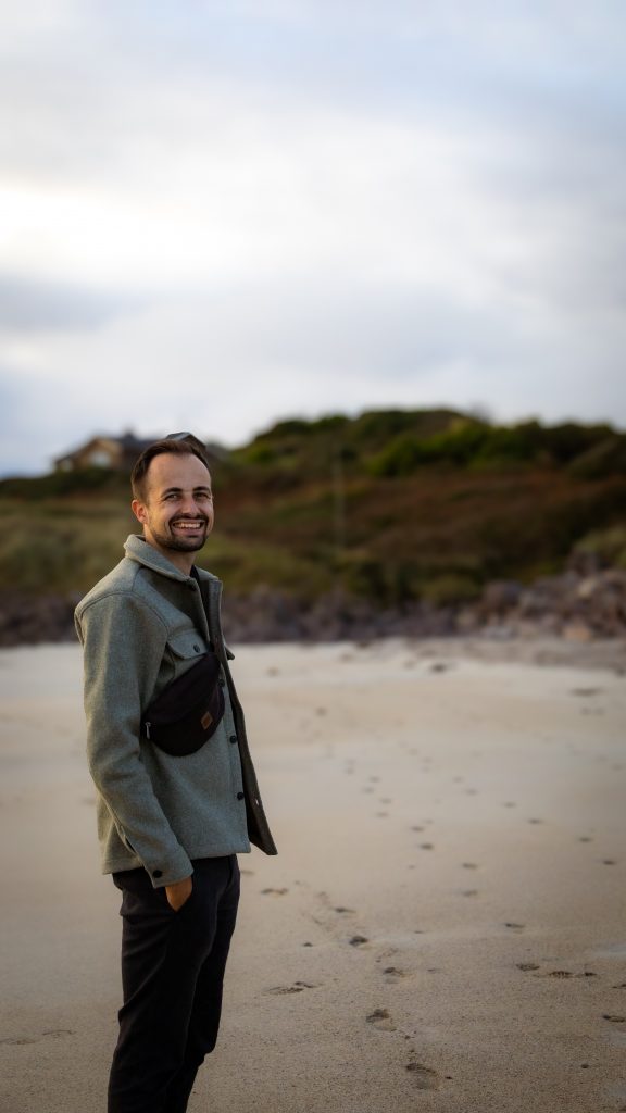 Philippe grinst an einem Strand mit grün, roten Büschen im Hintergrund an einem November Tag in Irland