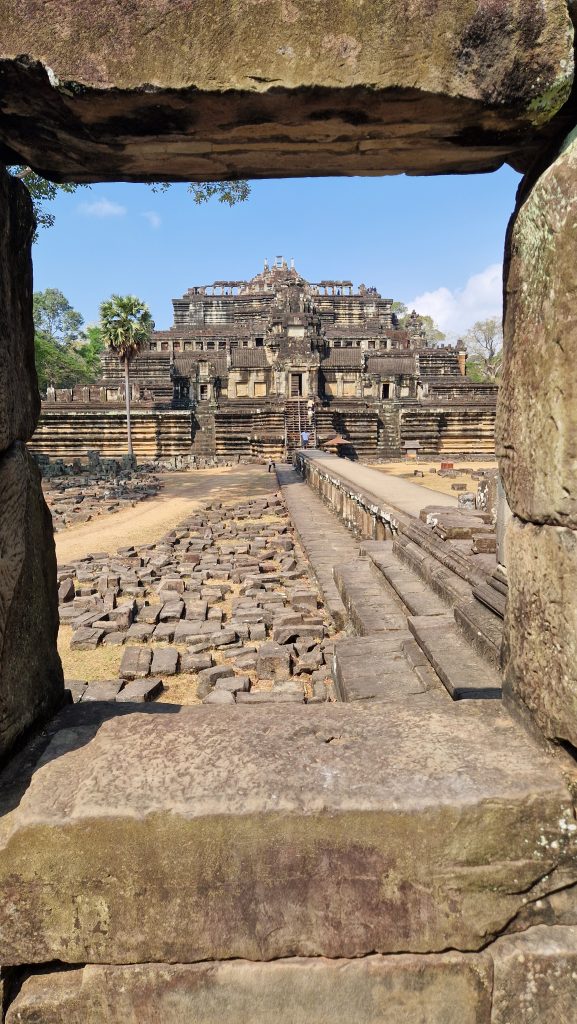Baphuon-Tempel Blick von Außen durch ein Steinfenster. Ein Steinsteg führt zu einem spitzen Tempel, der aus mehreren Terassen besteht, die durch steile Treppen verbunden sind.