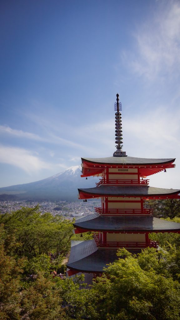 Die Chureito Pagoda ragt mit ihren geschwungenen Dächern aus grünen Bäumen hervor. Im Hintergrund sieht man den Fuji