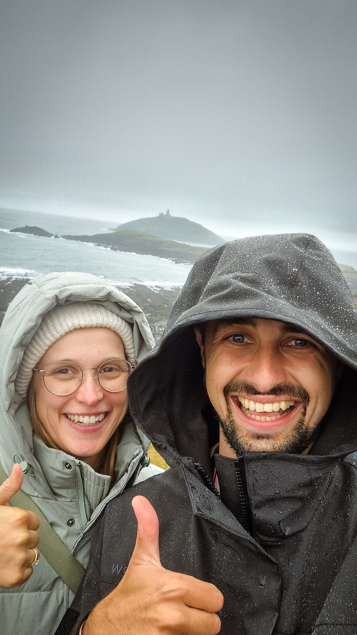 Lena und Philippe mit Regenklamotten im irischen Regen an der Küste. Lächelnd und Daumen hoch.