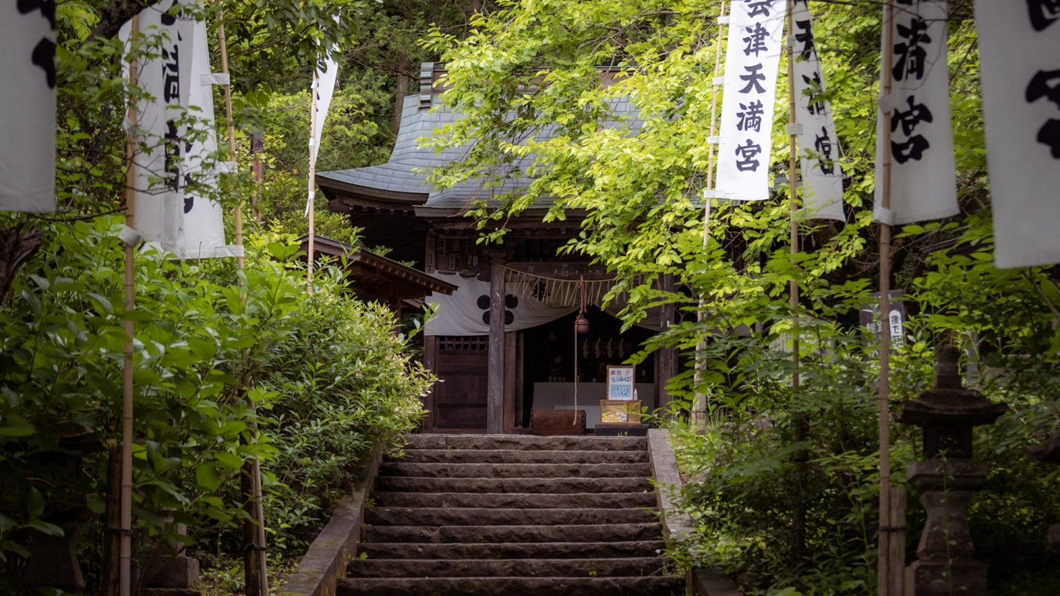 Eine Steintreppe führt gesäumt von grünen Bäumen und weißen Wimpeln mit japanischen Schriftzeichen zu einem Tempel.