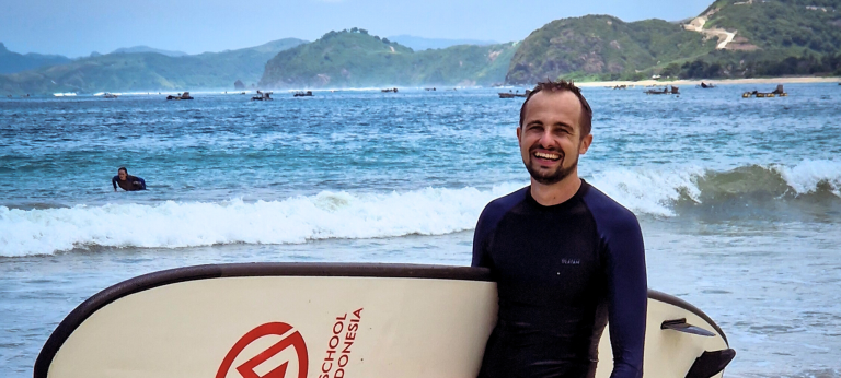 Philippe steht mit einem weißen Surfbrett unter dem Arm am Selong Belanak Beach. Im Hintergrund ist das blaue Meer und grüne Hügel der Insel Lombok zu sehen. Hey Welt Reisepodcast Lombok