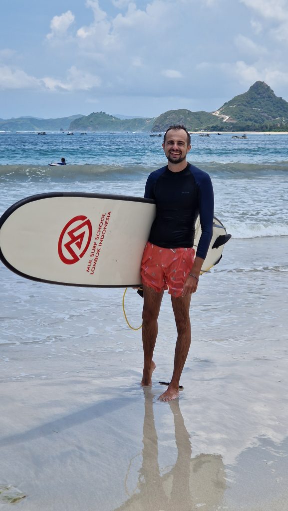 Philippe steht mit einem weißen Surfbrett unter dem Arm am Selong Belanak Beach. Im Hintergrund ist das blaue Meer und grüne Hügel der Insel Lombok zu sehen. 