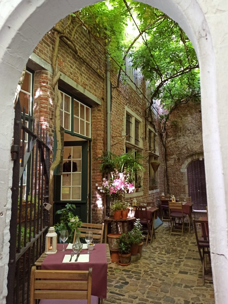Der Außenbereich eines kleinen Restaurants im Vlaeykensgang, ver steckten kleinen Gasse in Antwerpens zentrum. Man sieht alte Backsteinmauern und Efeu und kleine Tische mit Holzstühle.