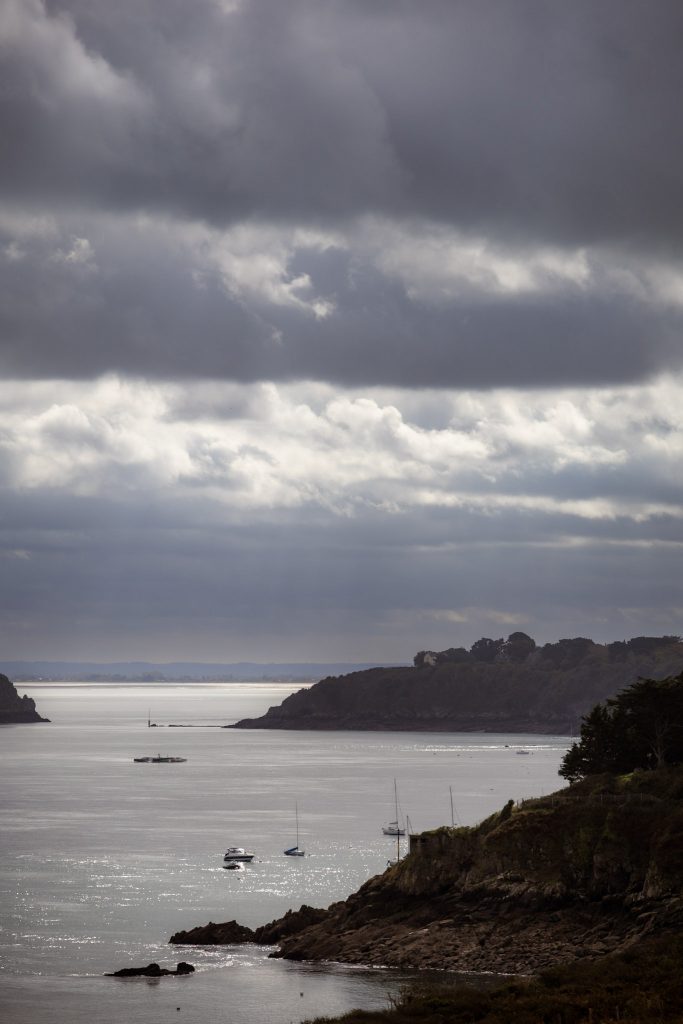 in den Wolken liegende Klippen an der Küste von Cancale, das Wasser glitzert von den leicht durchscheinenden Sonnenstrahlen und kleine Boote dümplen auf dem Meer