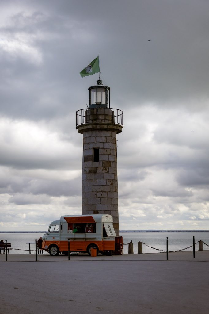 ein Stein-Leuchtturm fast allein am Pier in Cancale stehen, davor ein kleines Wein-Mobil, wo man ein Gläschen Wein kaufen kann
